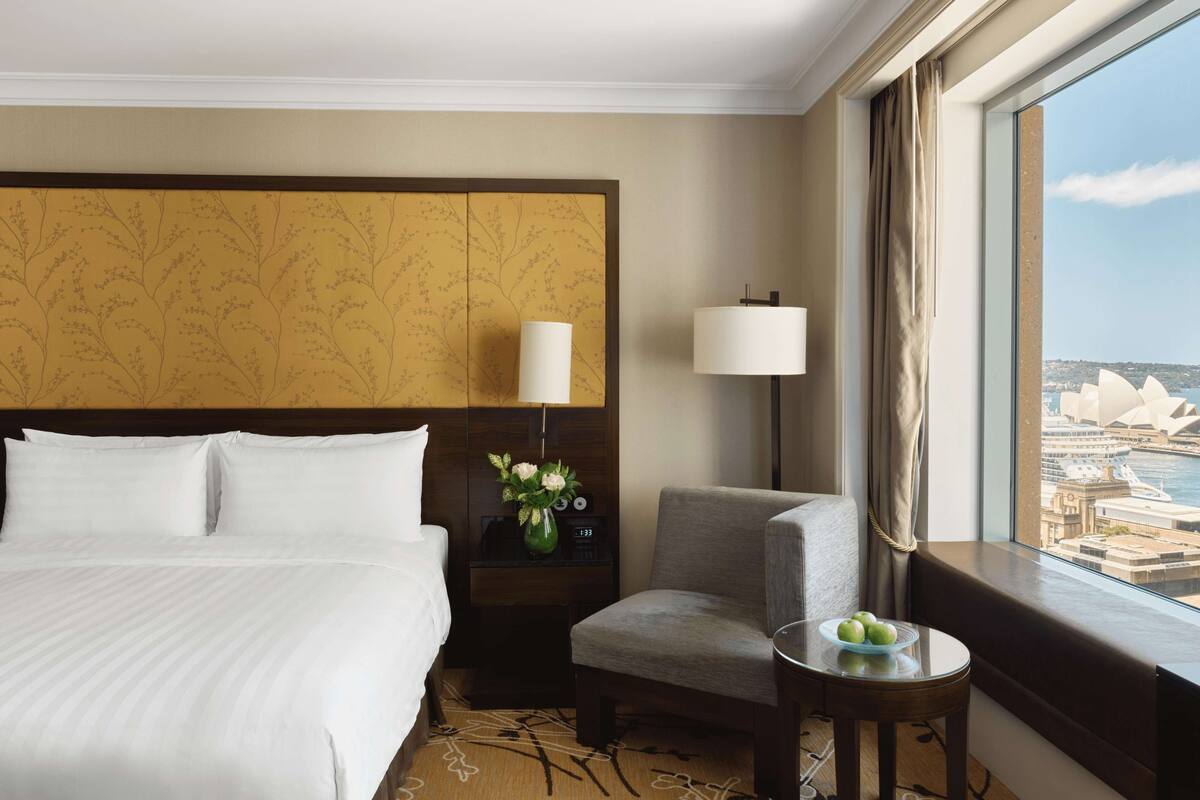 Ein Hotelzimmer mit einem großen Bett, einem Sessel, einem kleinen Tisch mit Früchten, einem Nachttisch mit Lampe und Blick auf ein bekanntes Wahrzeichen.