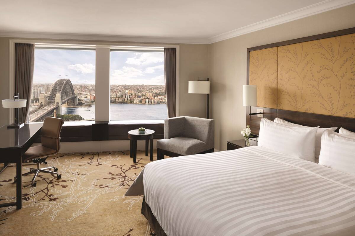A hotel room with a large bed, a desk, a chair, a small table, and a view of a cityscape and bridge.