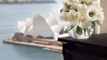 A piano with a vase of white flowers on a windowsill, overlooking a body of water and a distinctive white building.