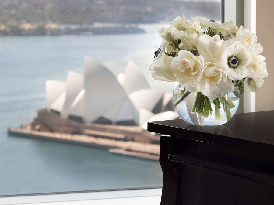 Ein Klavier mit einer Vase weißer Blumen auf einem Fensterbrett, mit Blick auf ein Gewässer und ein markantes weißes Gebäude.