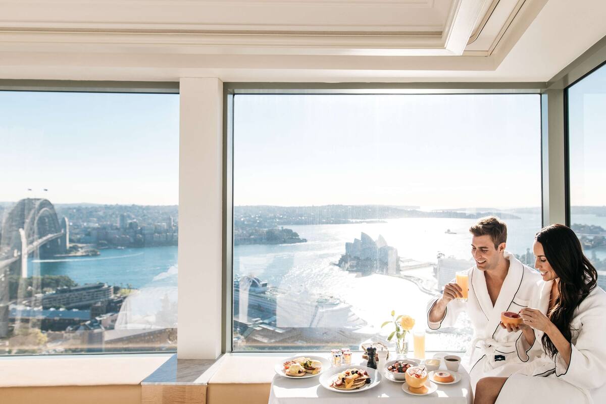 A couple in a hotel room with a large window overlooking a cityscape and a bridge.