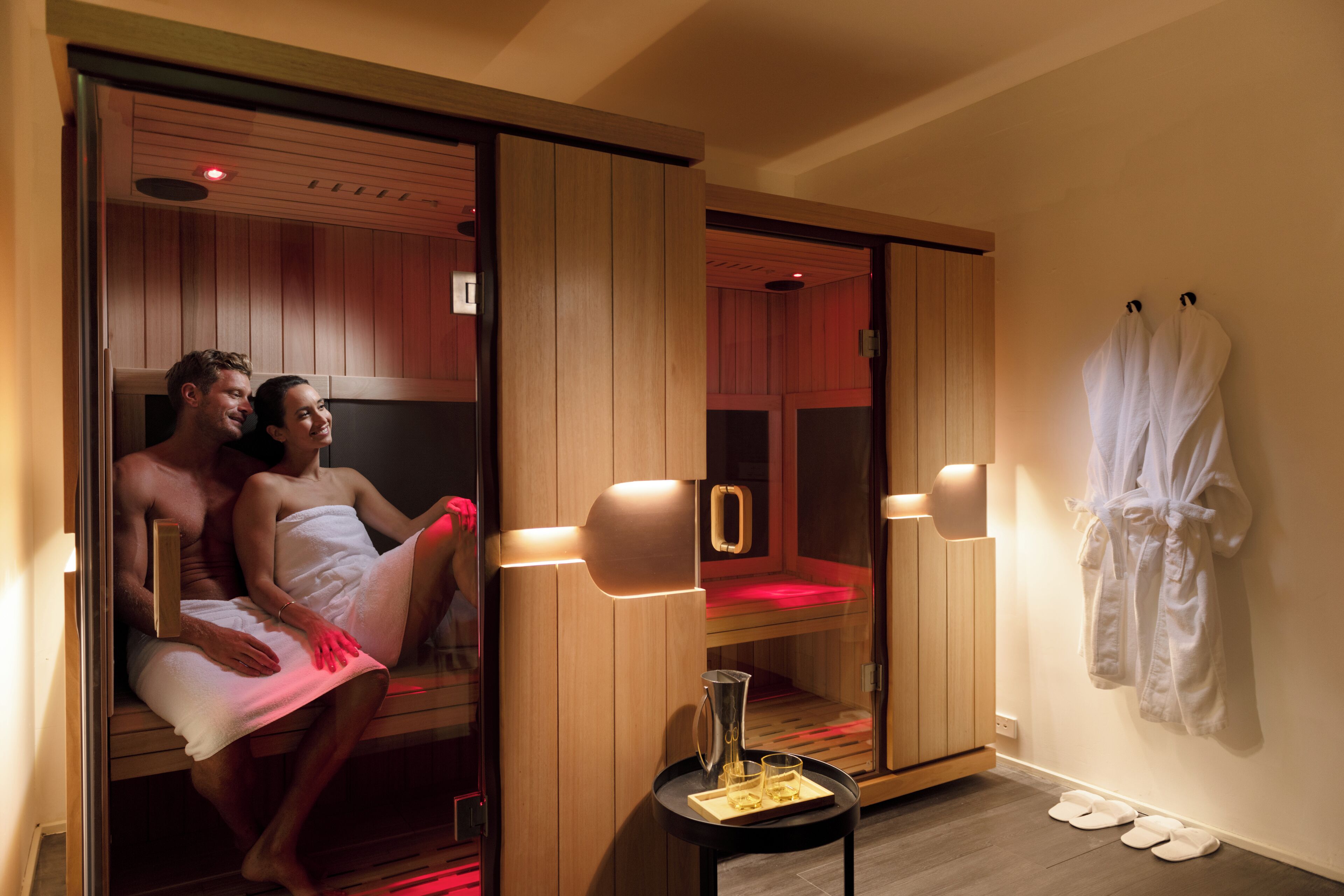 Una pareja en una sauna con paredes de madera y una puerta de vidrio, una pequeña mesa con una jarrón y vasos, y un par de zapatillas en el suelo.