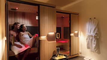 Una pareja en una sauna con paredes de madera y una puerta de vidrio, una pequeña mesa con una jarrón y vasos, y un par de zapatillas en el suelo.