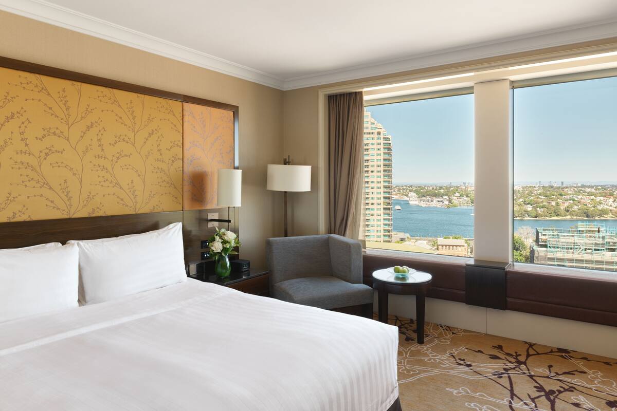 Ein Hotelzimmer mit einem großen Bett, einem Sessel, einem kleinen Tisch und Blick auf eine Stadtlandschaft und Wasser.