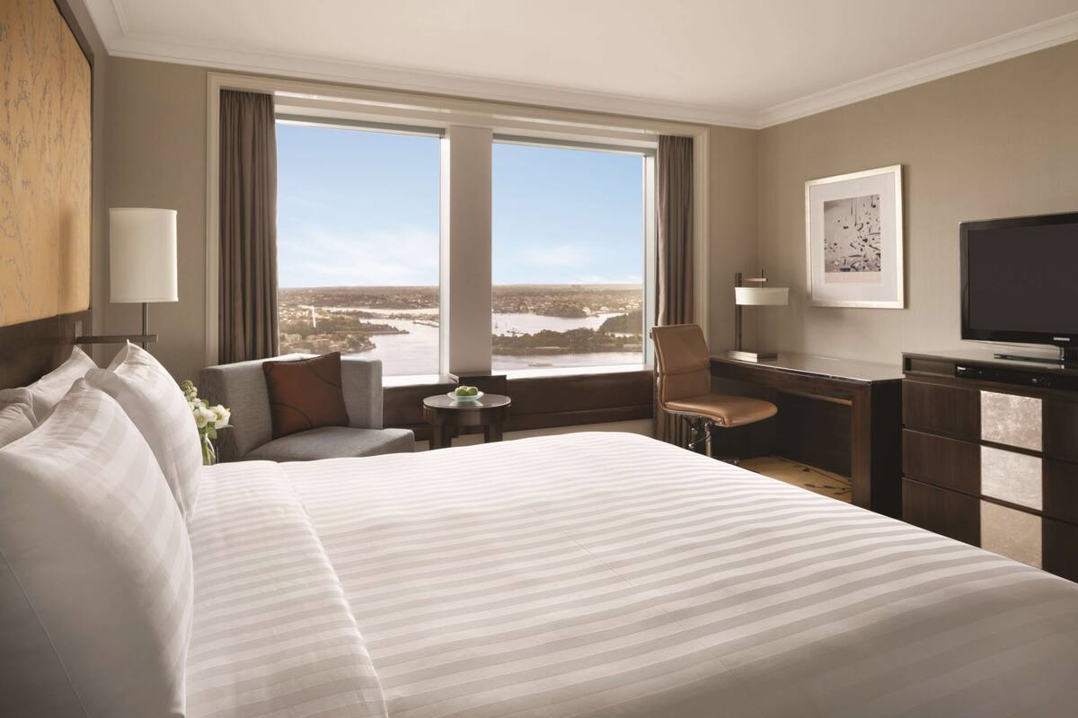 A hotel room with a large bed, a desk, a chair, and a view of a cityscape through large windows.