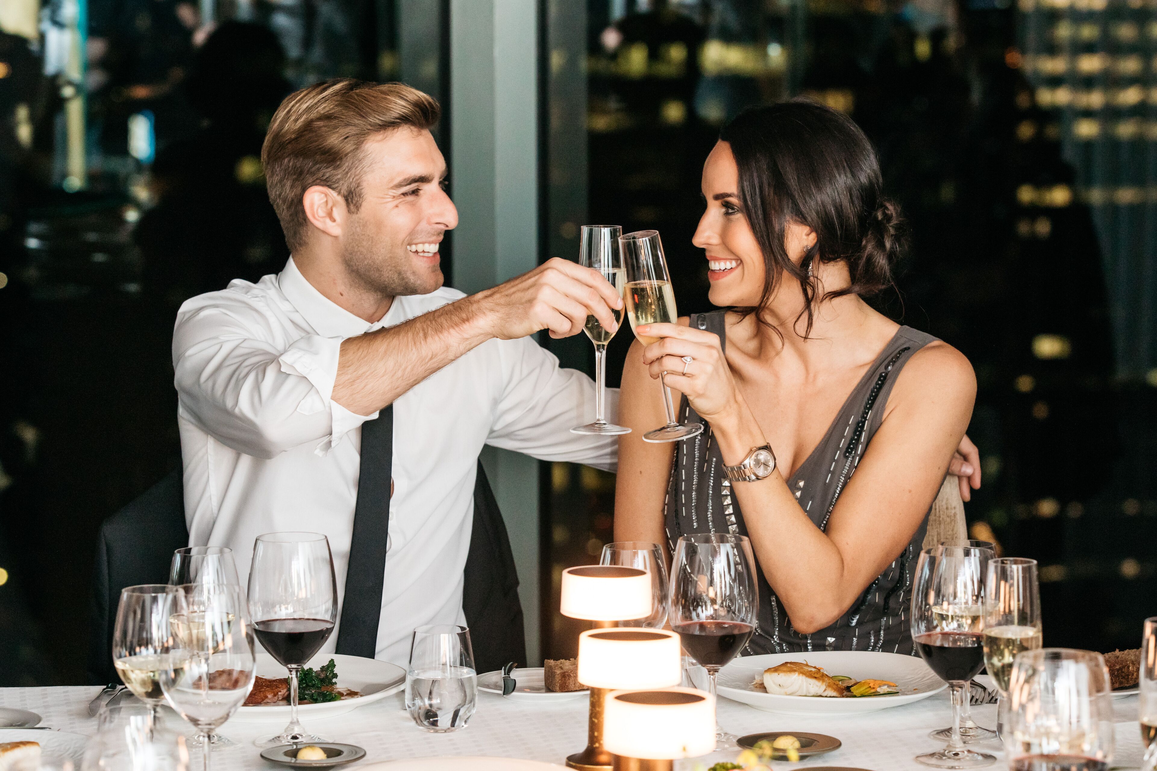Una coppia brinda con calici di champagne a un tavolo con diversi bicchieri da vino e piatti di cibo.