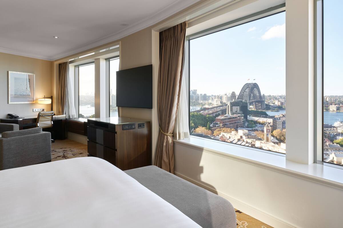 A hotel room with a large bed, a desk, a chair, a TV, and a view of a cityscape including a bridge.
