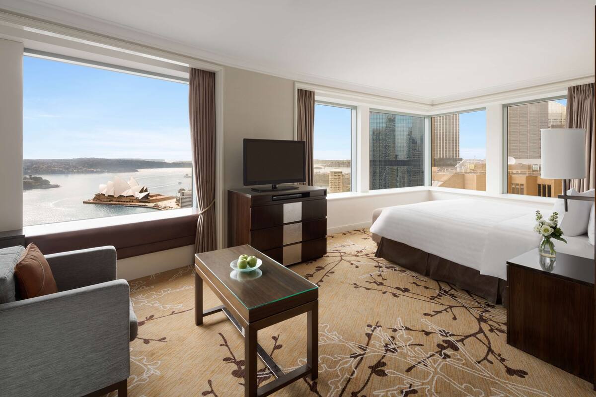 Ein Hotelzimmer mit einem großen Bett, einem Fernseher, einem kleinen Tisch mit einer Schüssel sowie Blick auf Stadt und Wasser.
