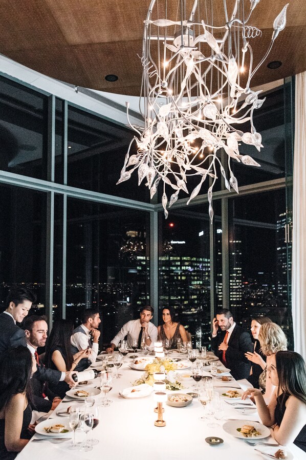 Un grupo de personas cenando en una mesa con vistas a la ciudad, un candelabro moderno y un mantel blanco.