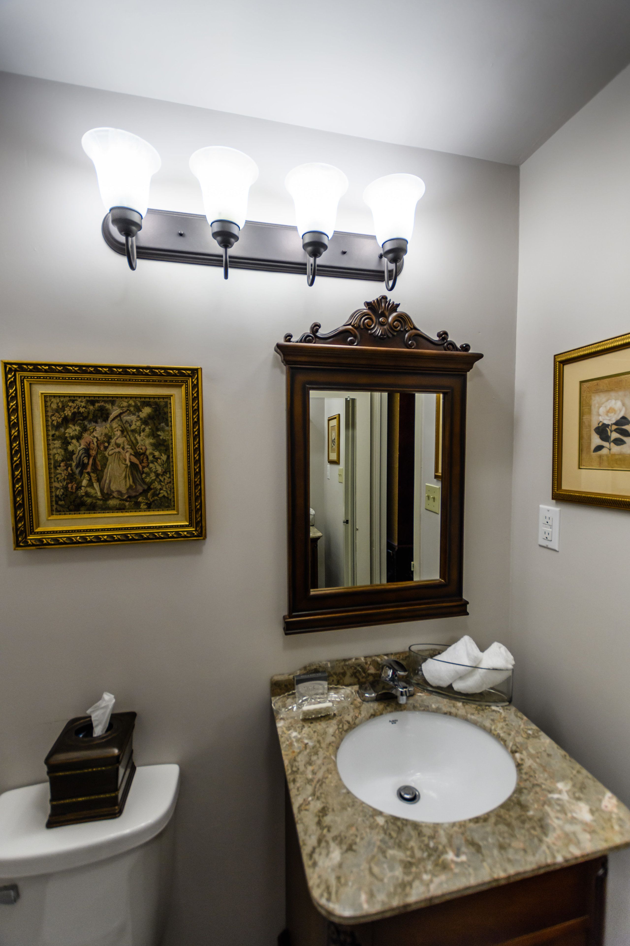 Un baño con inodoro, lavabo y espejo de tocador. Hay luces empotradas sobre el espejo y cuadros en las paredes.