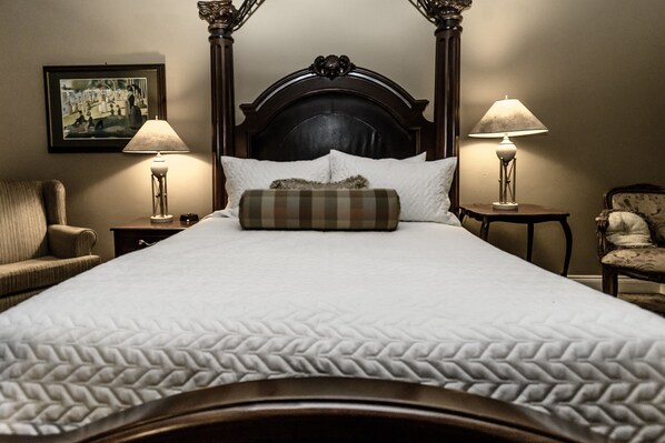 Een netjes opgemaakt bed met een gewatteerde deken, omringd door twee nachtkastjes met lampen, een stoel en een ingelijst schilderij aan de muur.