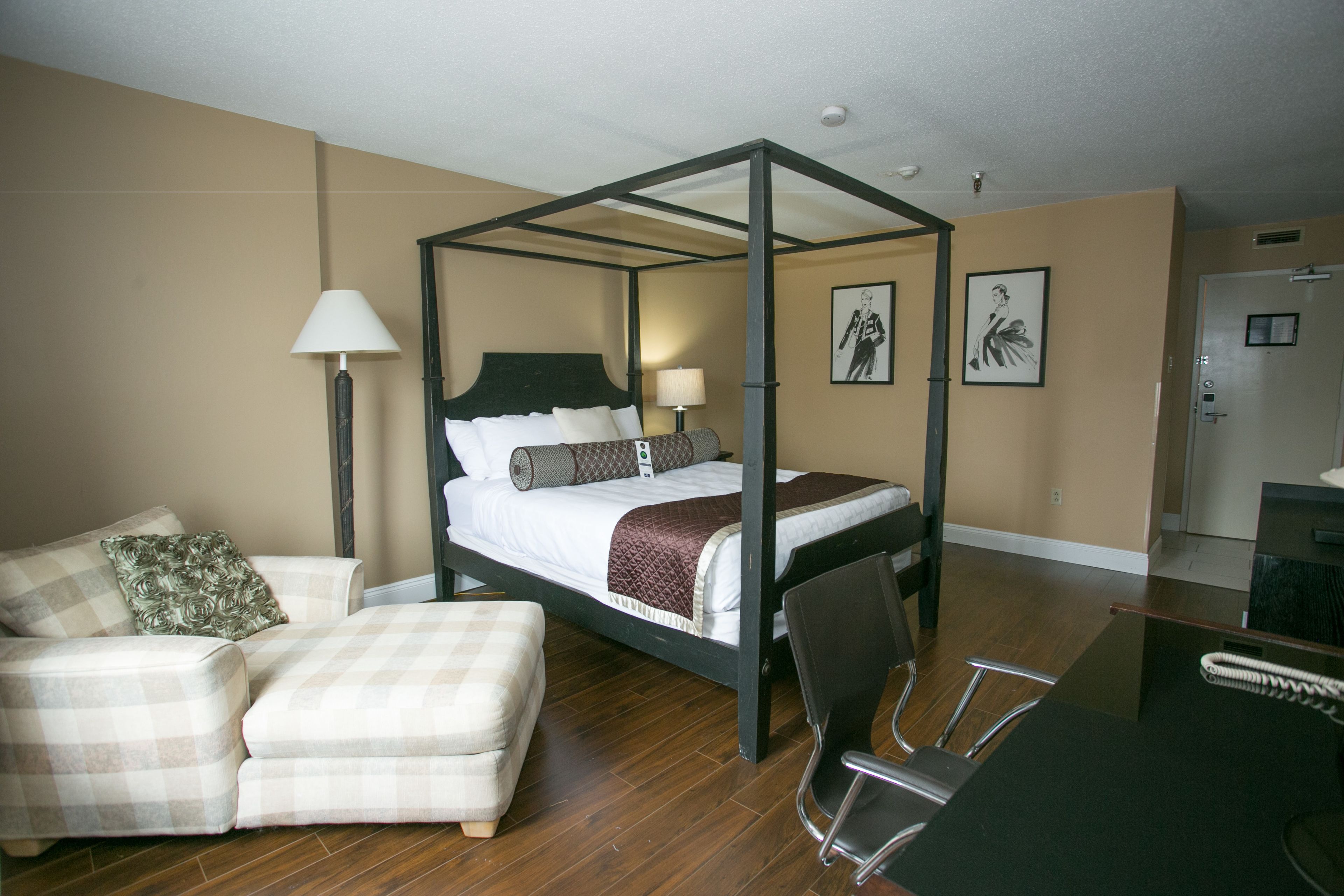 A four-poster bed with a wooden frame, a bedside table with a lamp, a chair, a sofa, and a door in the background.