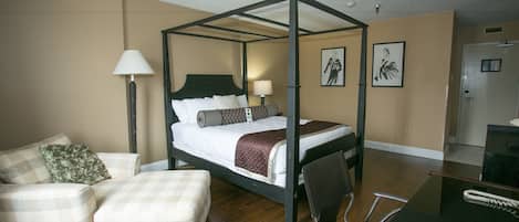 A four-poster bed with a wooden frame, a bedside table with a lamp, a chair, a sofa, and a door in the background.