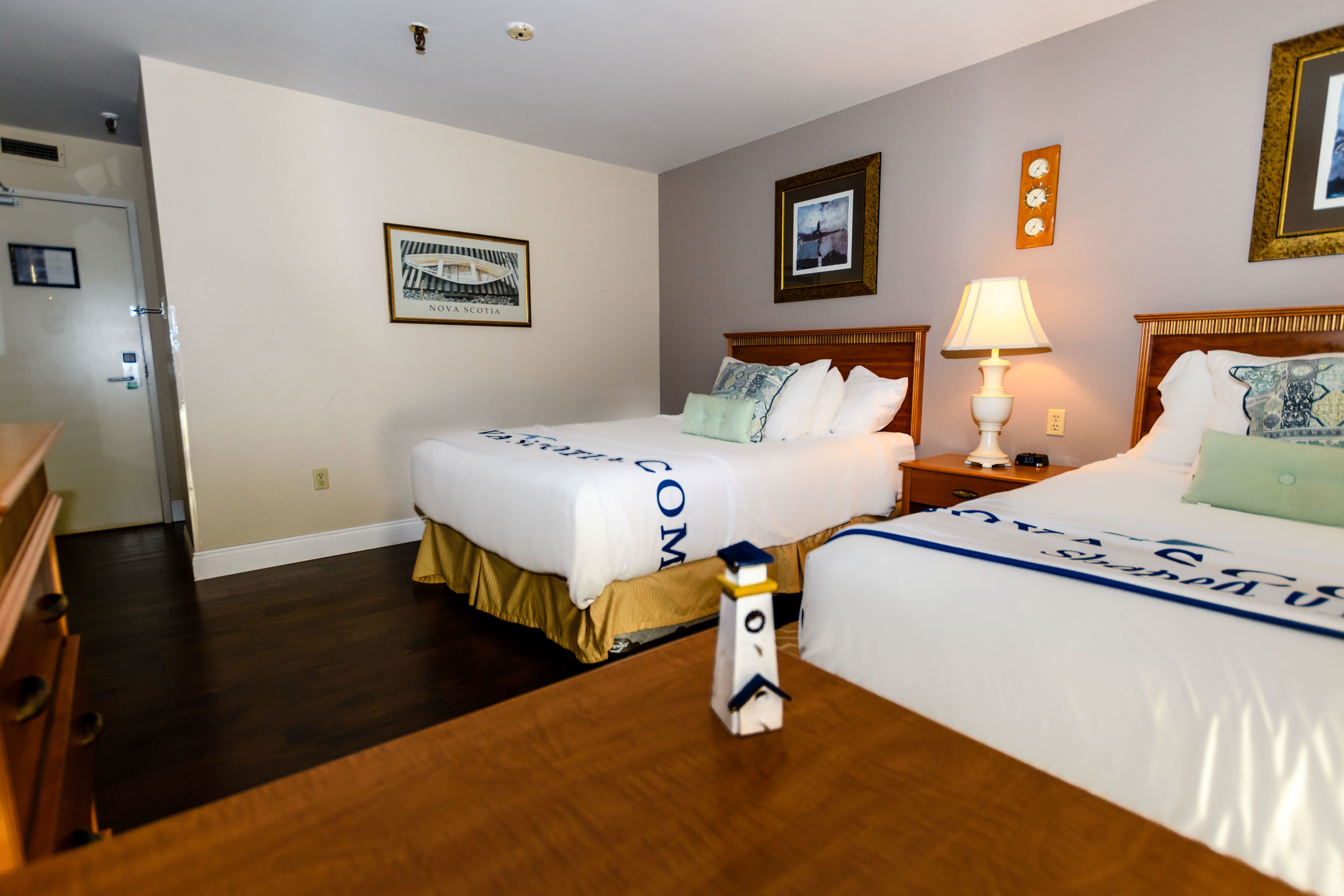 Une chambre d’hôtel avec deux lits, une commode en bois, une table de chevet avec une lampe et un tableau encadré au mur.