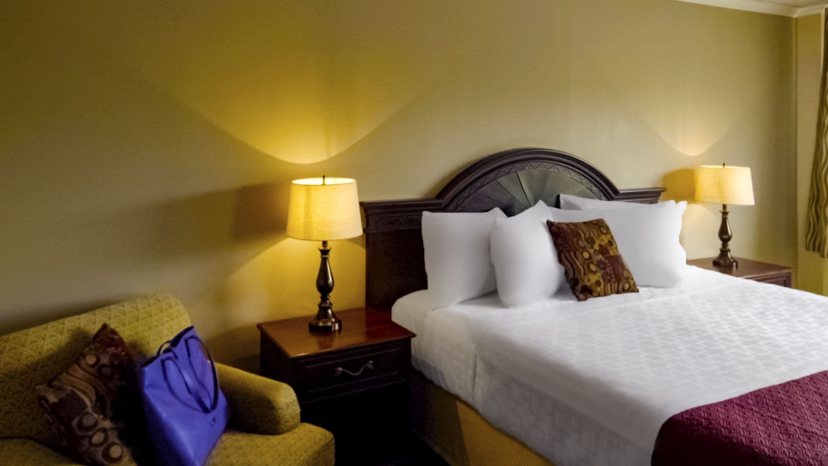 A hotel room with a large bed, two bedside lamps, a nightstand, a chair, and a patterned throw blanket.