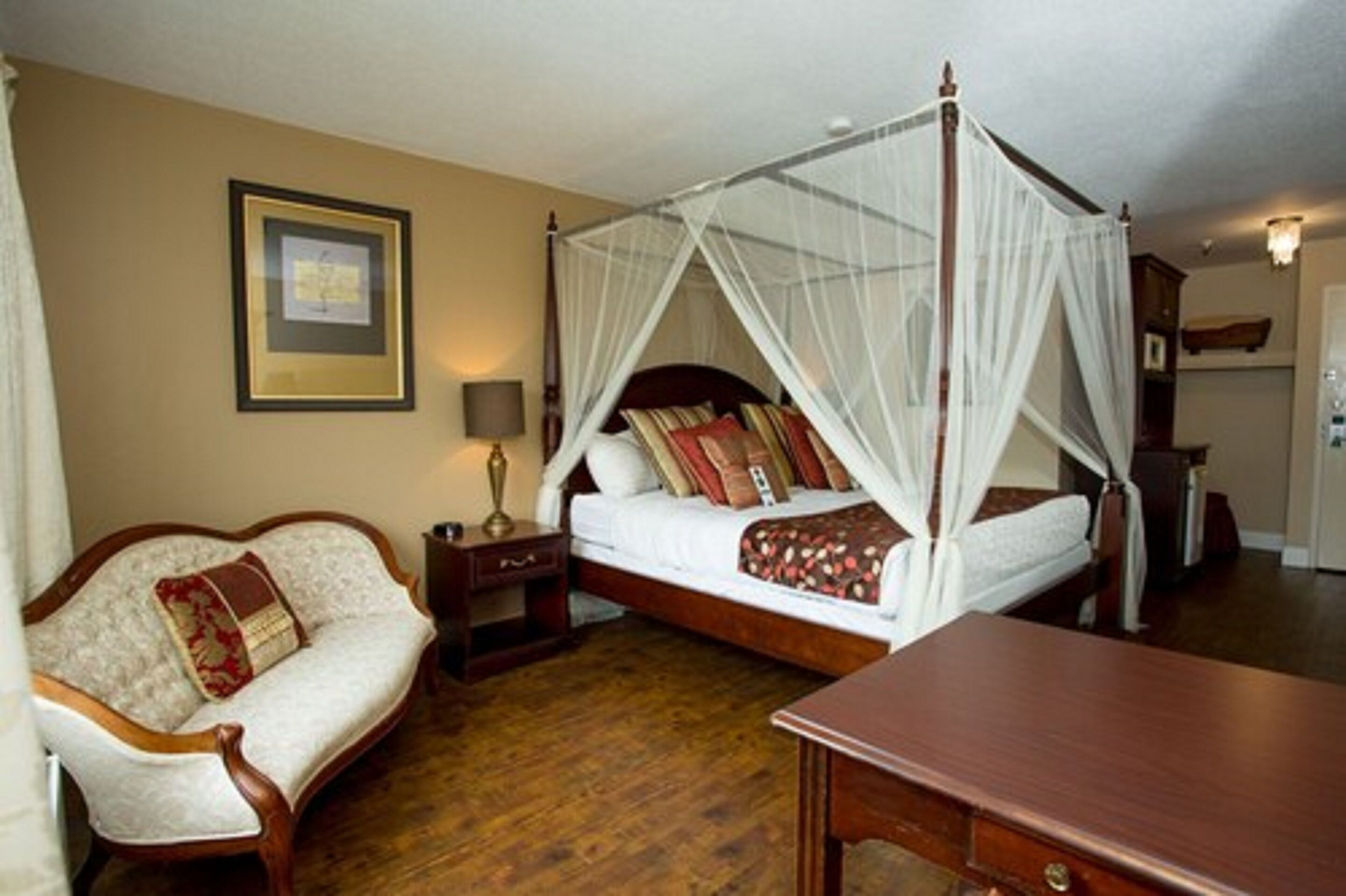 A four-poster canopy bed with a nightstand and lamp, a sofa with a decorative pillow, and a wooden table in a hotel room.