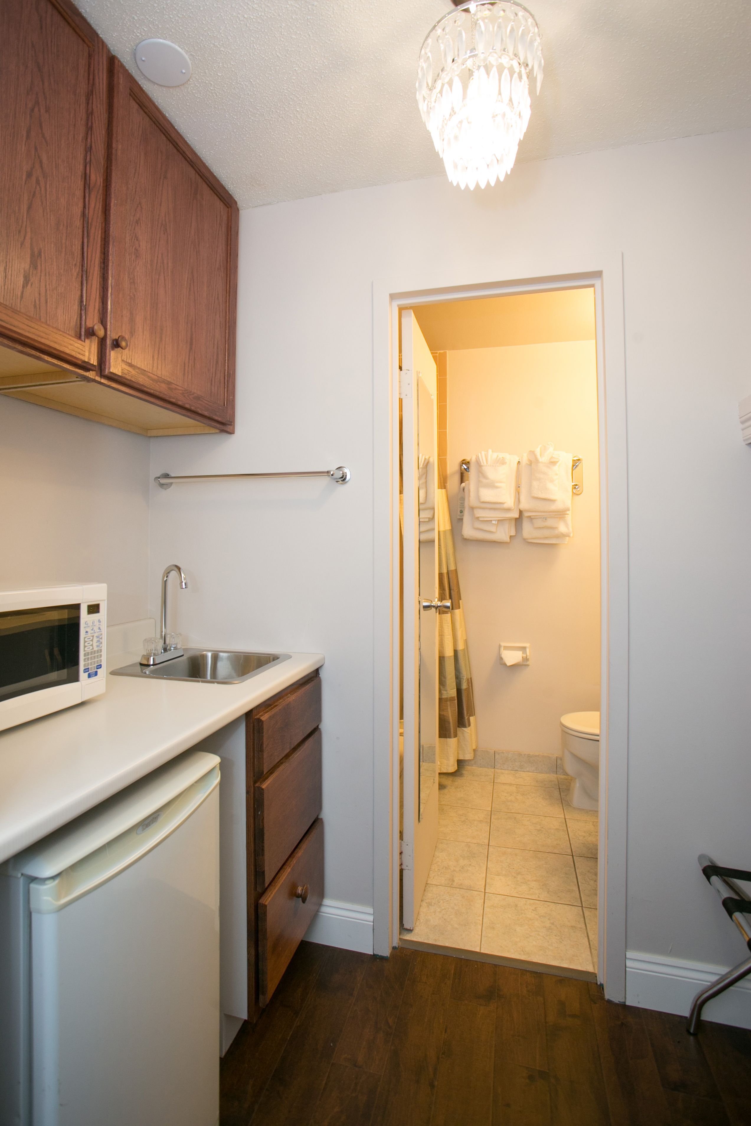 A compact kitchen with a microwave, sink, and wooden cabinets. A chandelier hangs from the ceiling, and a towel rack is mounted on the wall.