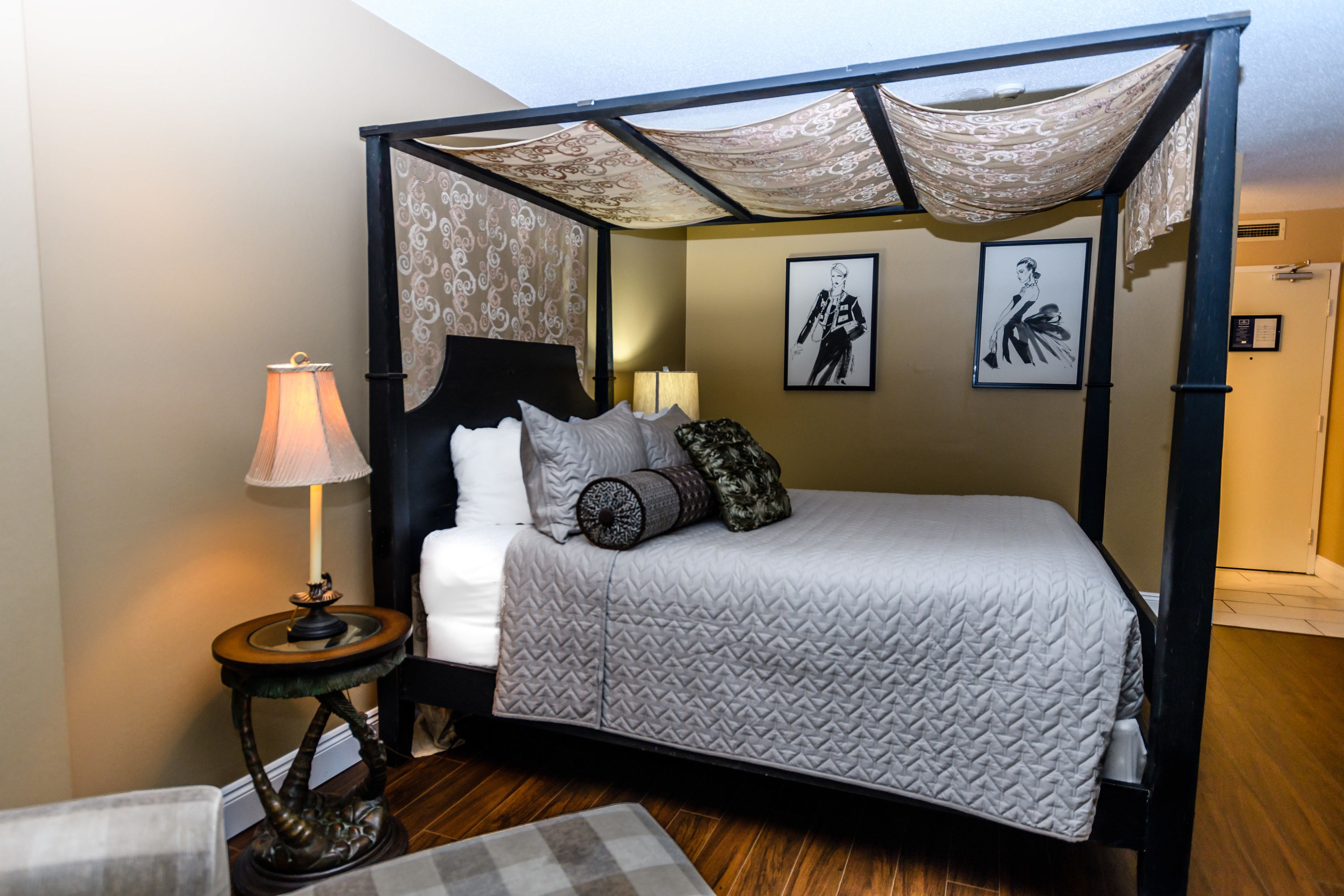 A four-poster bed with a quilted bedspread, two bedside lamps, and a small round table with a lamp.