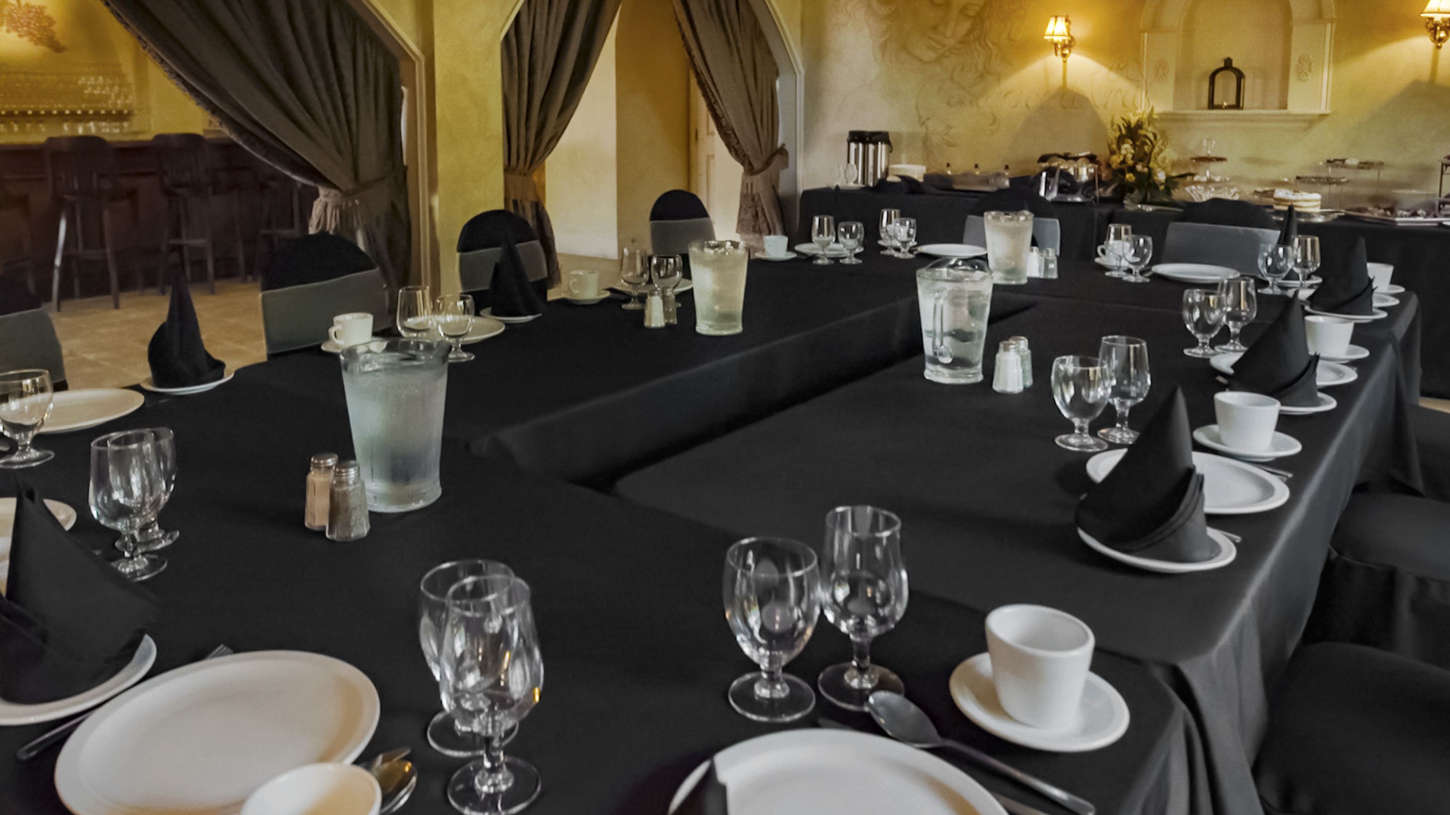 A formal dining room with long tables set for a meal, featuring black tablecloths, white plates, and clear glassware.