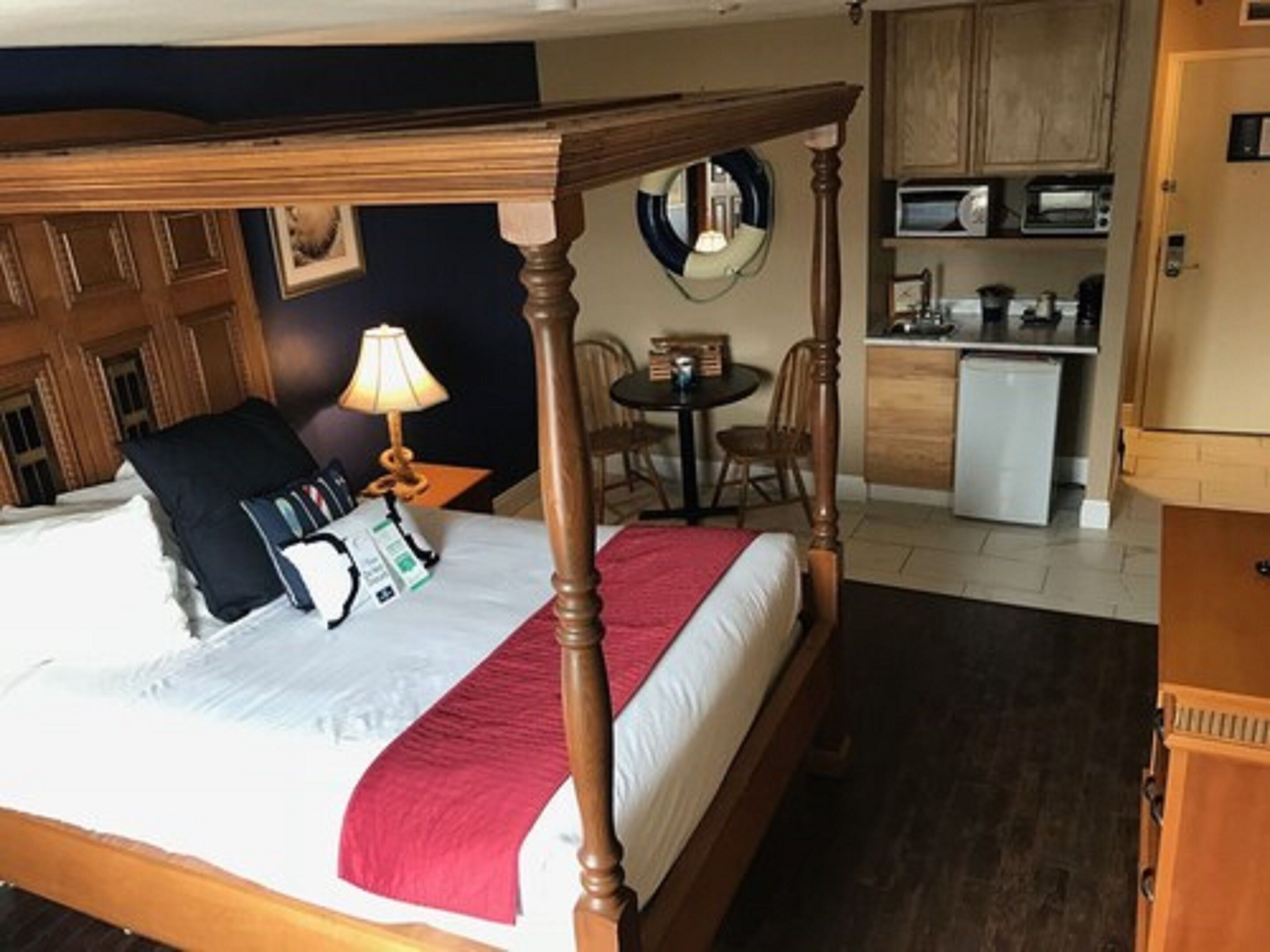 Un lit à baldaquin avec un couvre-lit rouge, une table de chevet avec une lampe, une petite table ronde avec des chaises, et, au fond, une petite cuisine.