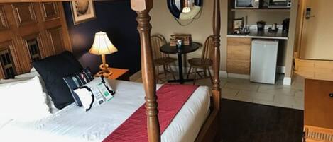 Una cama con dosel con una manta roja, una mesita de noche con lámpara, una pequeña mesa redonda con sillas y una kitchenette al fondo.