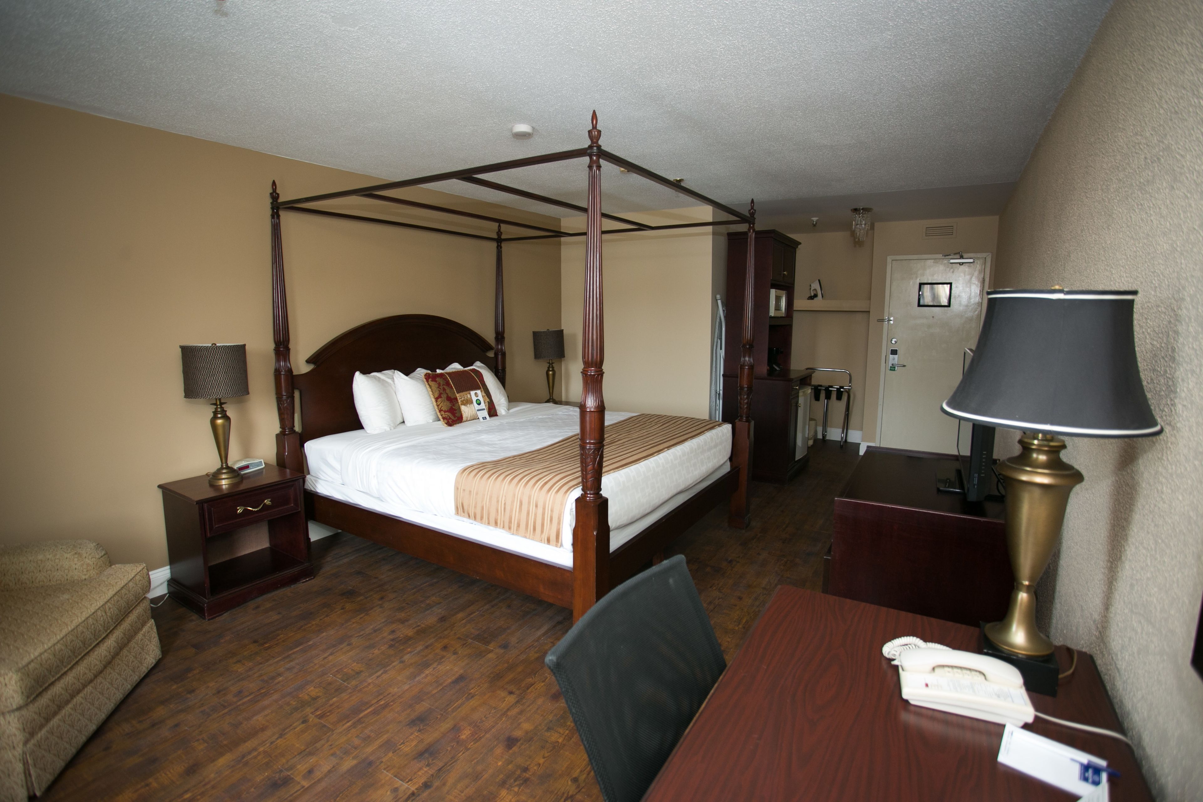 A four-poster bed with a wooden frame, a bedside table with a lamp, a chair, and a desk with a telephone.