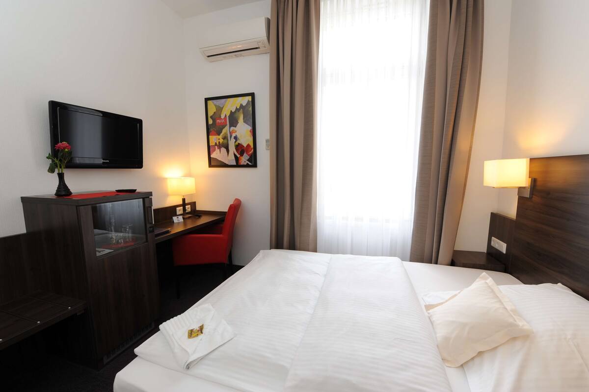 Quarto de hotel com cama, escrivaninha, cadeira, televisão e um quadro na parede.