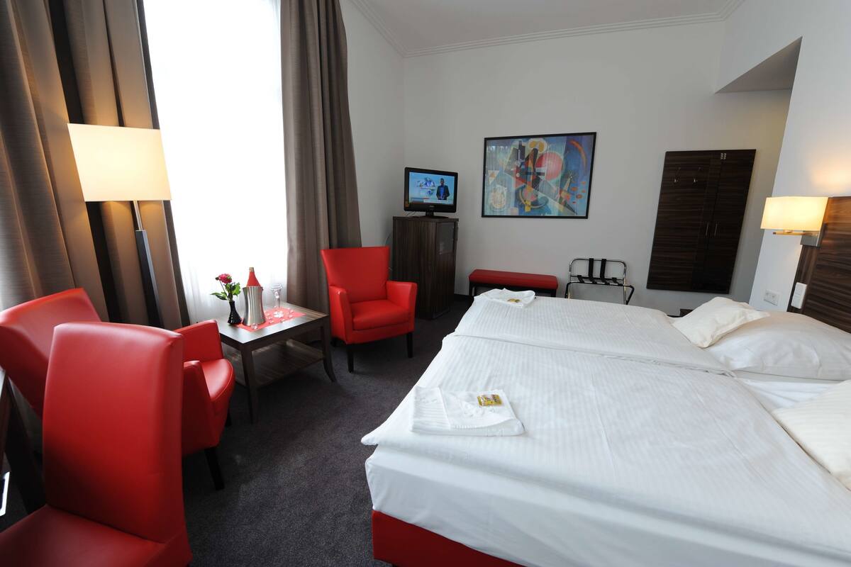 Quarto de hotel com uma cama grande, duas cadeiras vermelhas, uma mesinha com abajur, uma televisão e um quadro na parede.