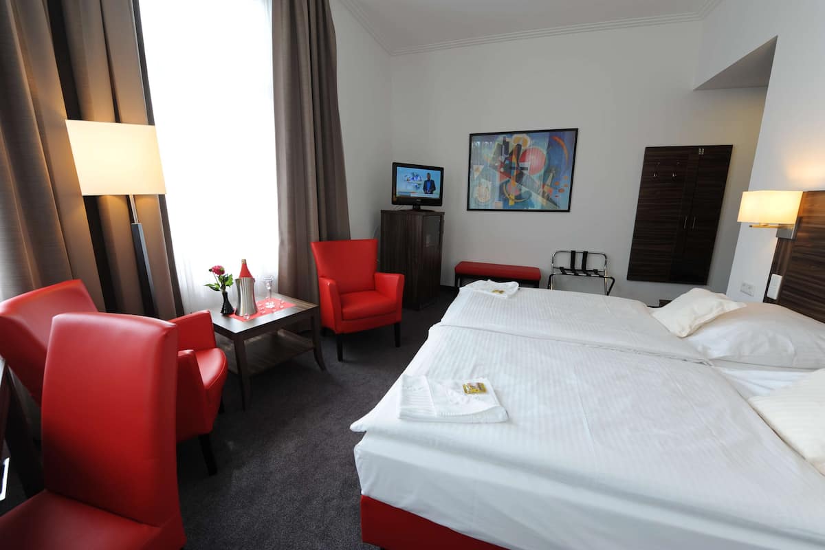 Quarto de hotel com uma cama grande, duas cadeiras vermelhas, uma escrivaninha com abajur, televisão e um quadro na parede.