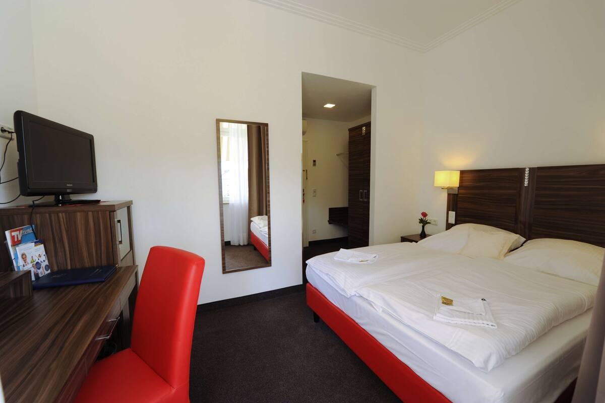 Quarto de hotel com cama, escrivaninha com televisão, uma cadeira vermelha e espelho.