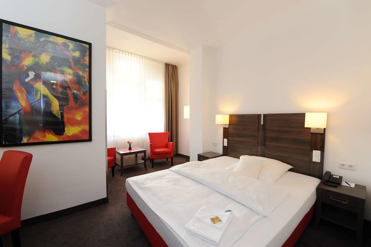 Quarto de hotel com uma cama grande, duas mesas de cabeceira, uma escrivaninha, uma cadeira e um quadro na parede.