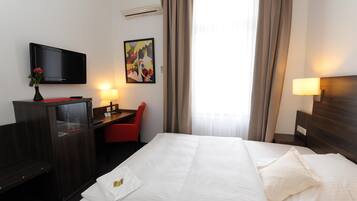 Una habitación de hotel con cama, escritorio, silla, televisor, obras de arte y una ventana con cortinas.