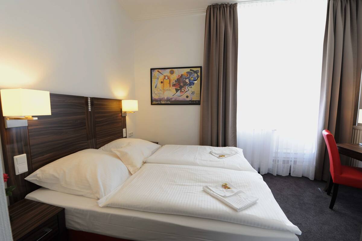 Quarto de hotel com cama, duas luminárias de cabeceira, um quadro na parede, janela com cortinas e uma cadeira vermelha.