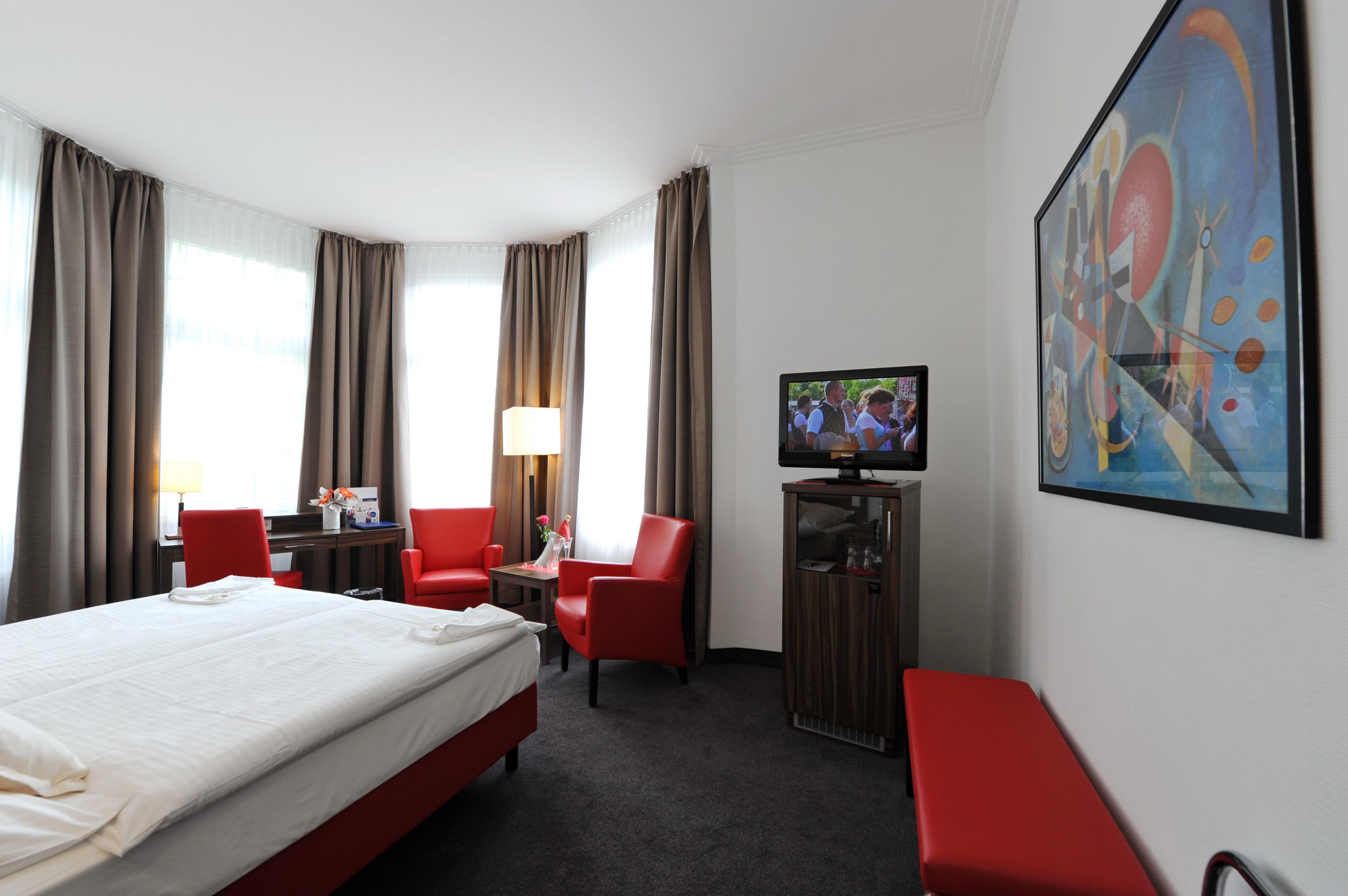 A hotel room with a large bed, two red chairs, a desk with a lamp, a television, and a painting on the wall.