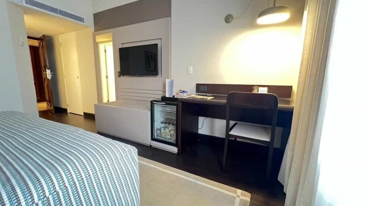 Habitación de hotel pequeña y funcional con cama, kitchenette, escritorio con silla y televisor montado en la pared.