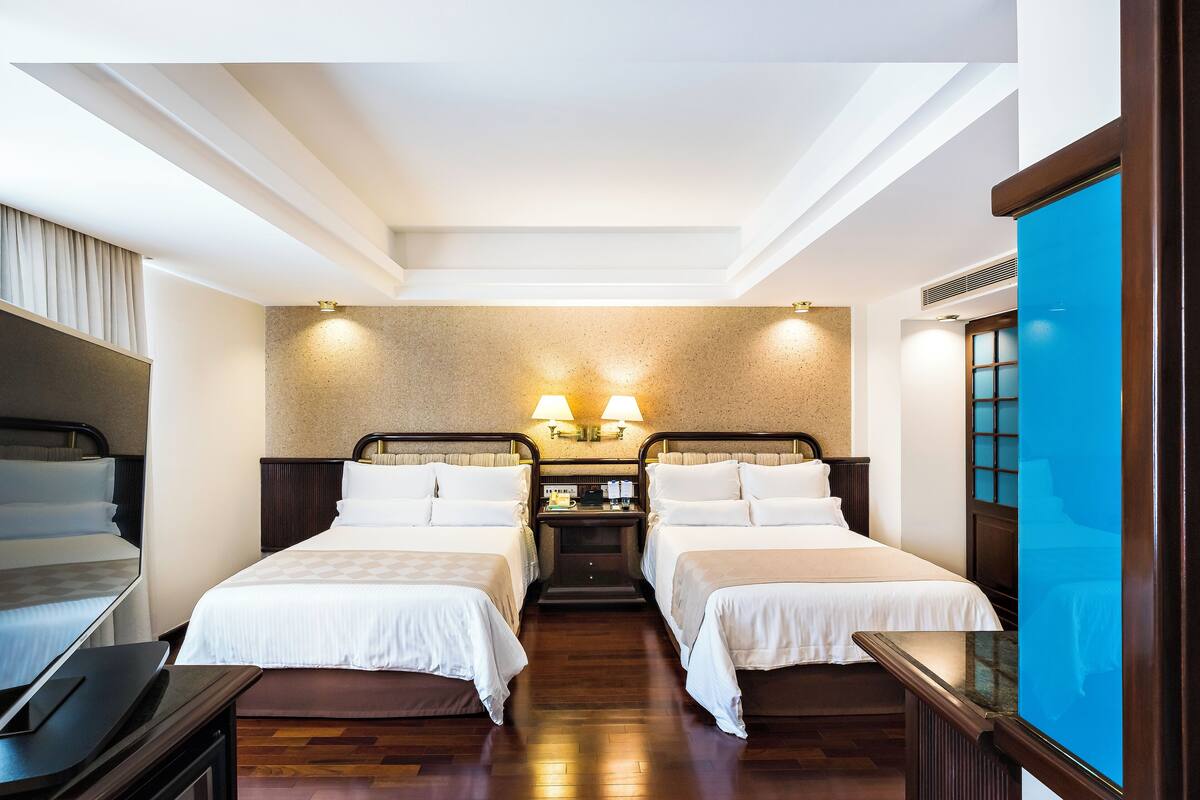 Habitación de hotel con dos camas individuales, una mesita pequeña, una mesita de noche y un ventanal grande.