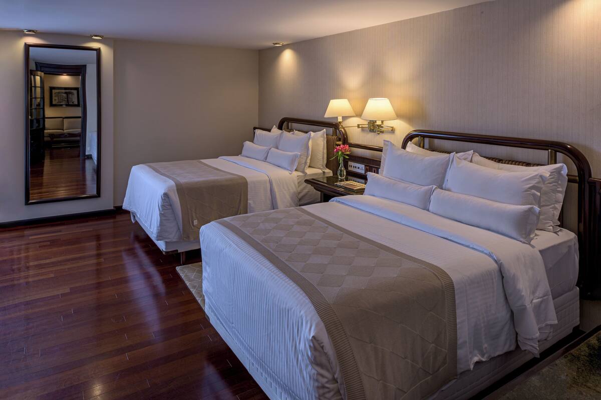 Habitación de hotel con dos camas, una mesita de noche con lámparas, un espejo y suelo de madera.
