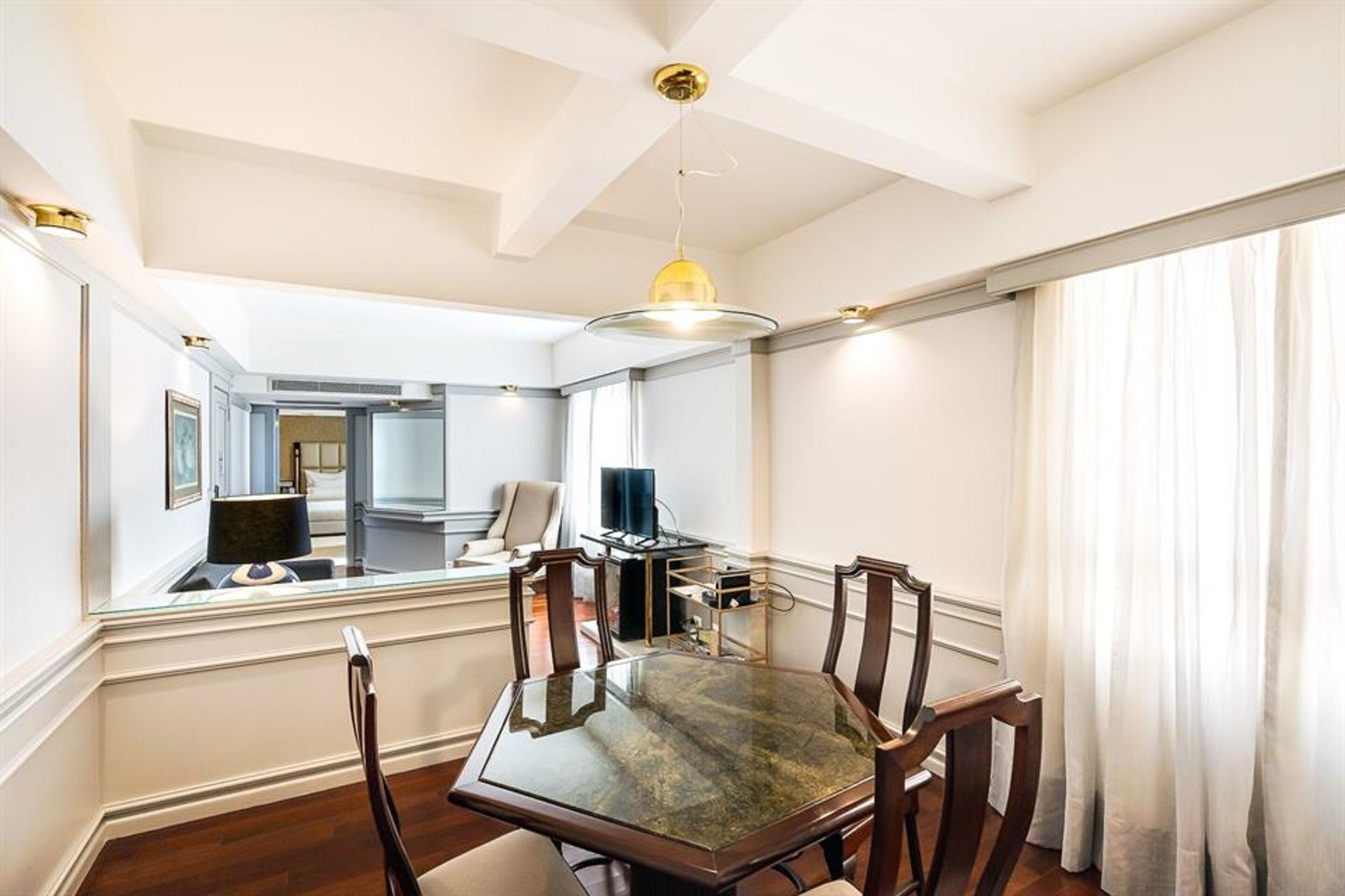 A dining area with a glass-top table, wooden chairs, a pendant light, and a view of a living room through a glass partition.
