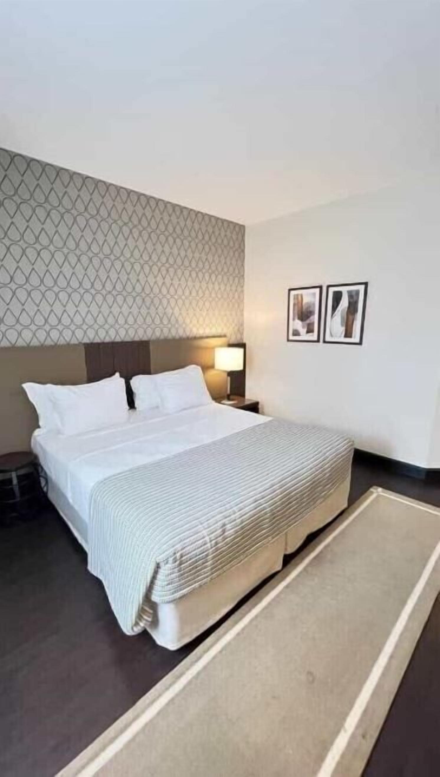 Una habitació d'hotel amb un llit, una tauleta de nit amb una làmpada, un mur amb un disseny decoratiu i dues fotografies emmarcades a la paret.