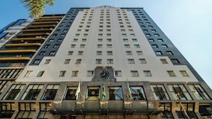 A tall, modern building with a clock at the center, multiple windows, and flags at the entrance.