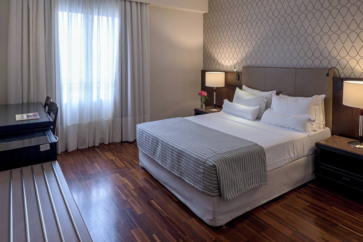 Una habitación de hotel con cama, mesitas de noche, escritorio y ventana con cortinas.