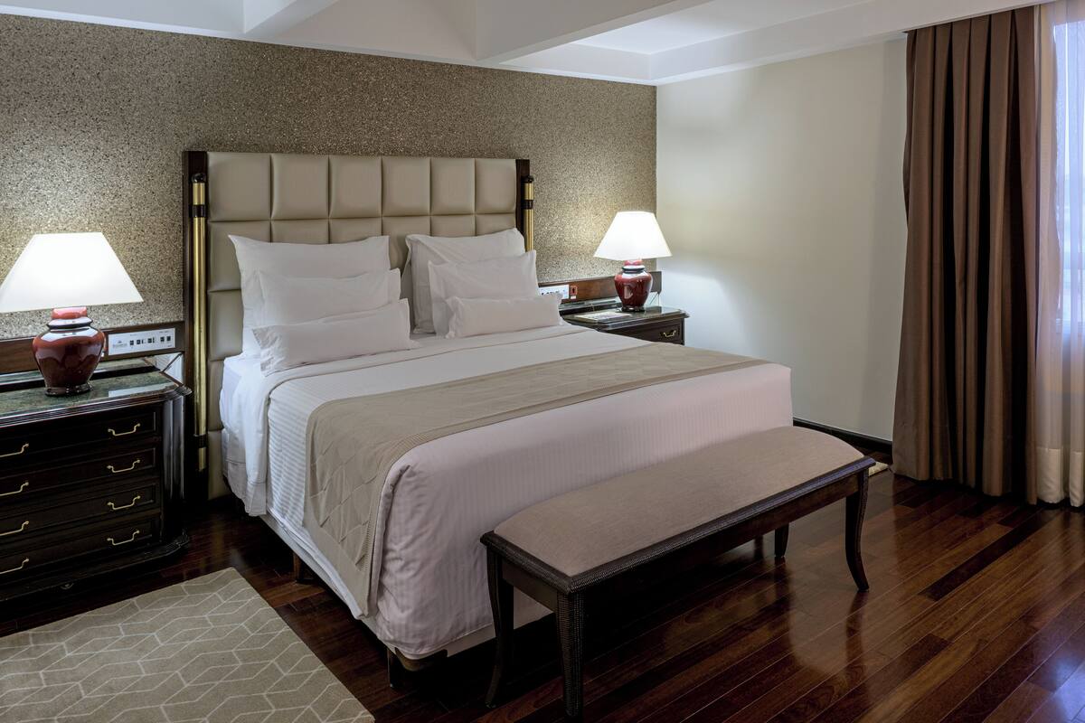 Habitación de hotel con una cama grande, dos mesitas de noche con lámparas, un banco, un aparador y vistas al exterior a través de cortinas translúcidas.