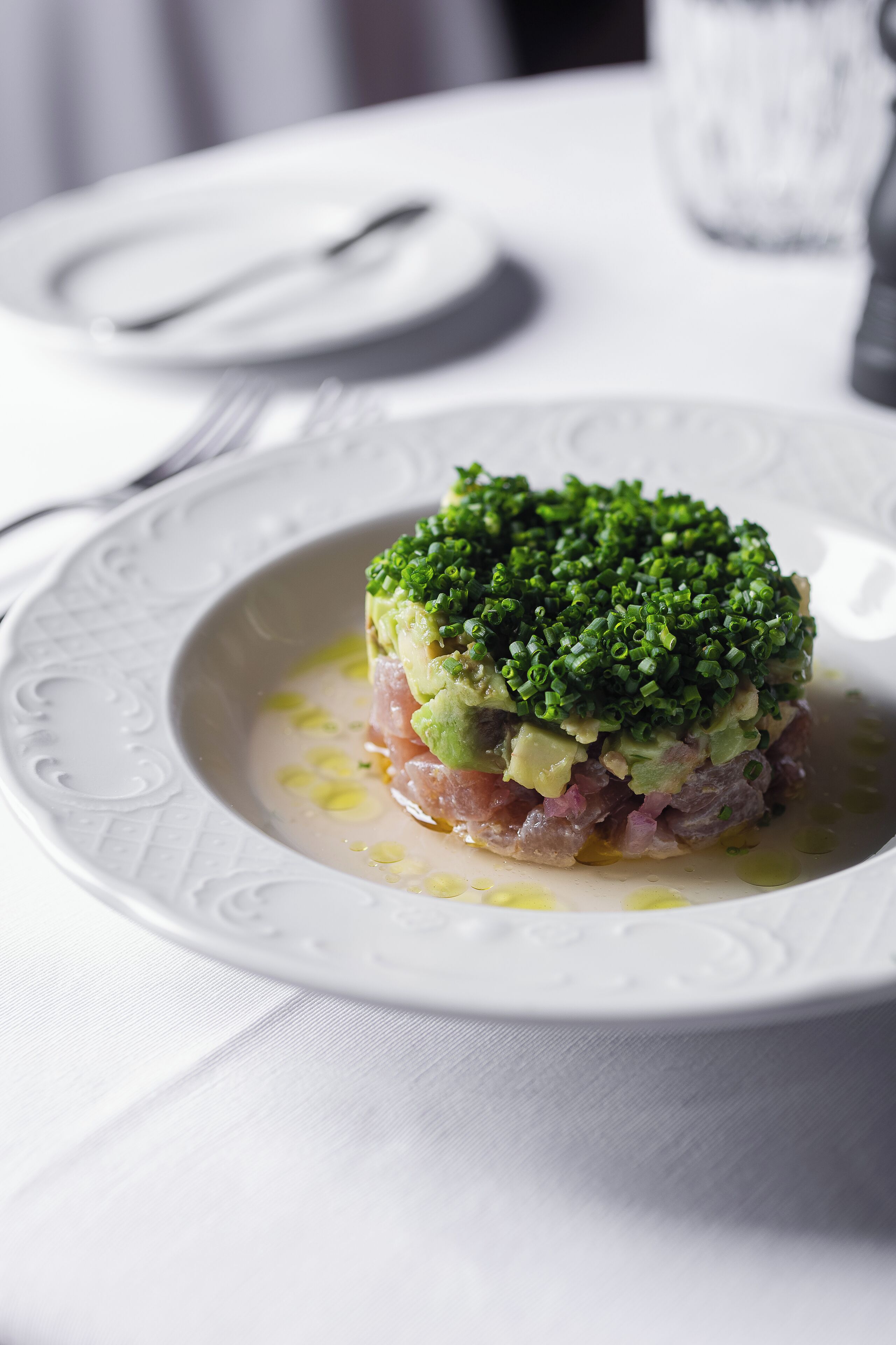 A white plate with a layered dish, possibly a seafood tartare, garnished with green onions, on a white tablecloth.