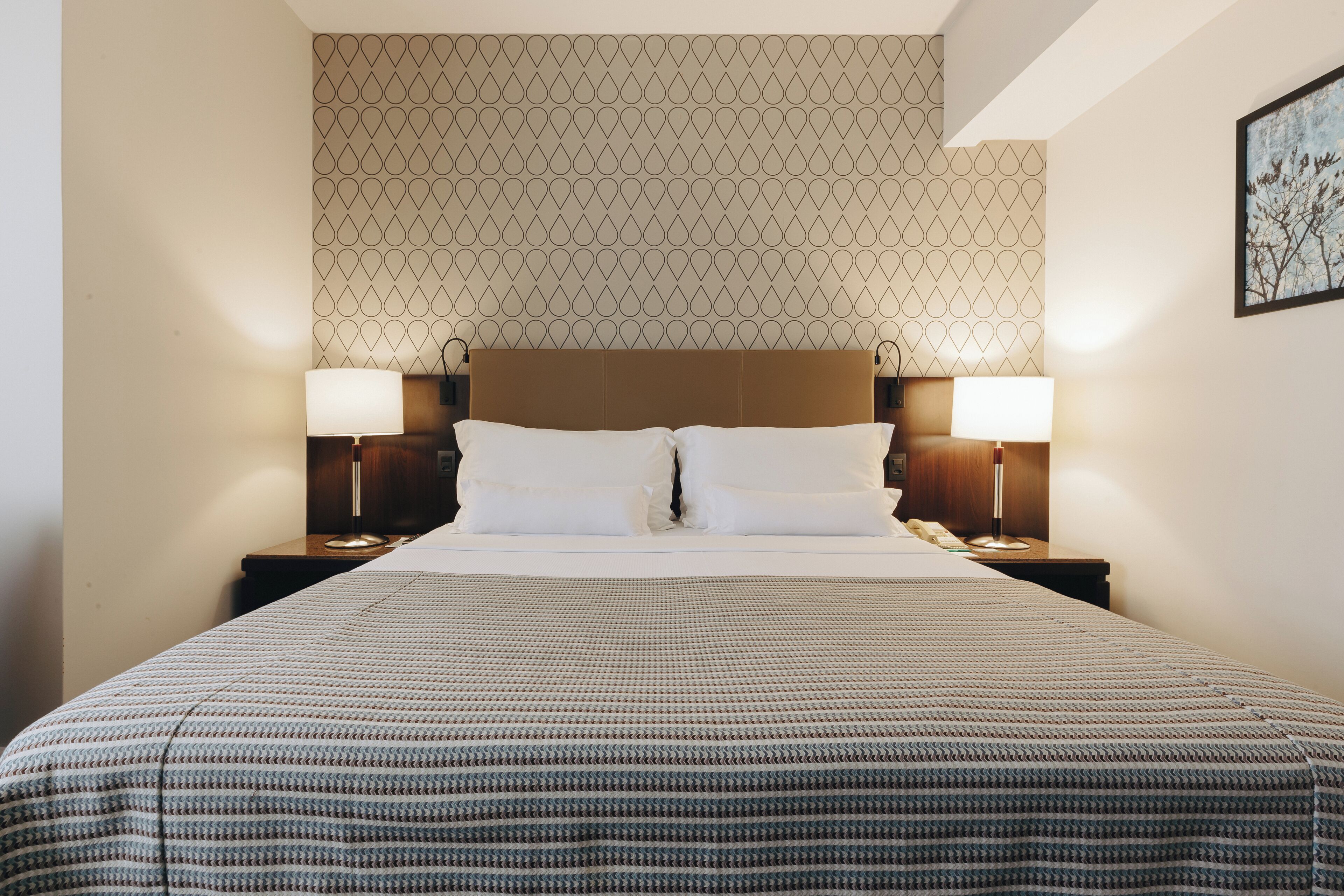 A neatly made bed with a patterned bedspread, two bedside lamps, and a patterned wallpaper on the wall behind.