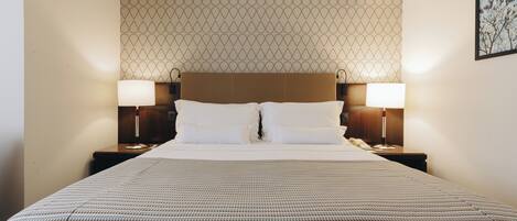A neatly made bed with a patterned bedspread, two bedside lamps, and a patterned wallpaper on the wall behind.