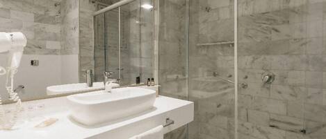 Un baño moderno con lavamanos blanco, dos espejos, cabina de ducha con mampara de vidrio y paredes de mármol.