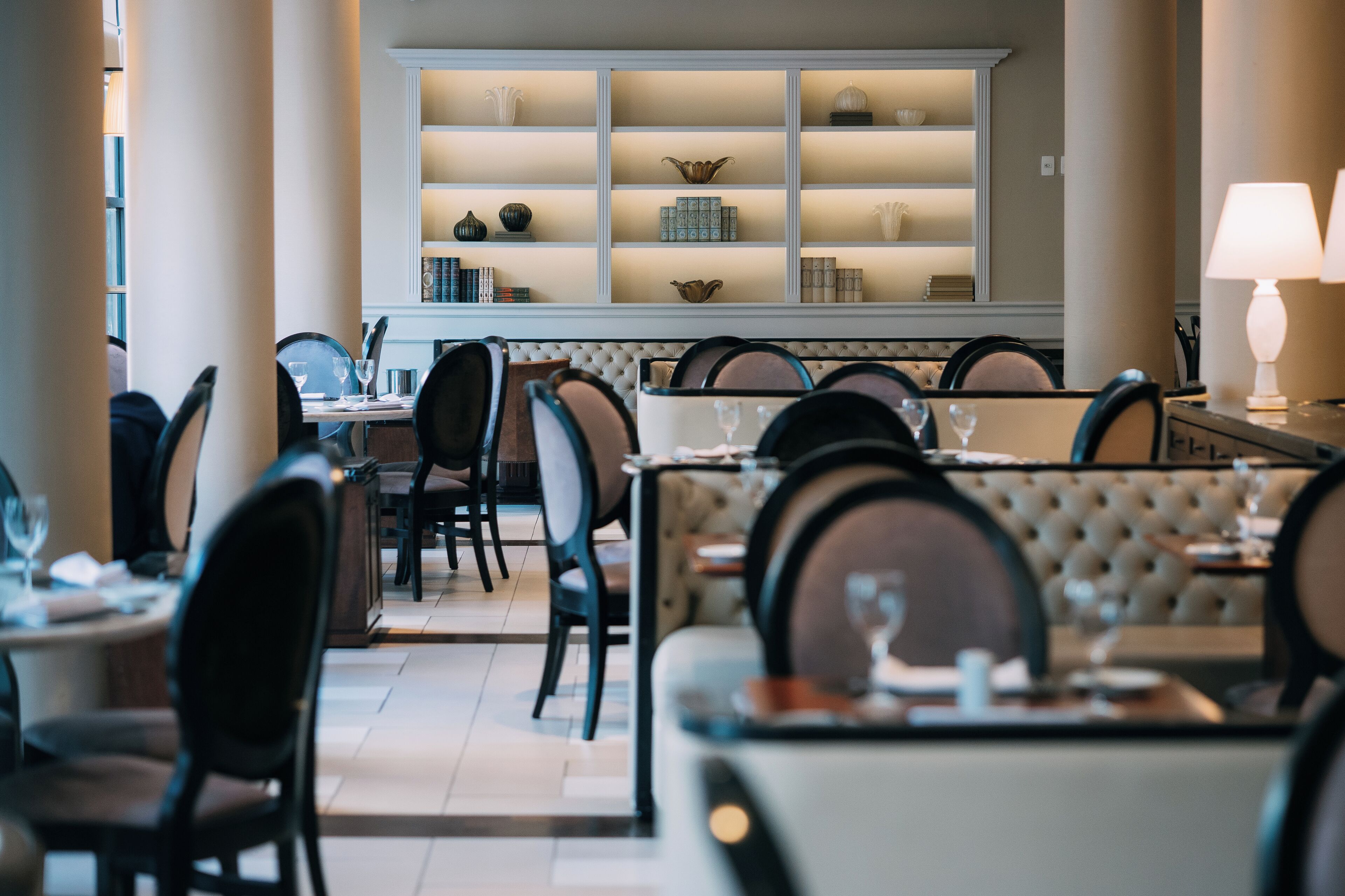 Een restaurant met een lange tafel, stoelen en een muur met planken waarop diverse objecten staan.