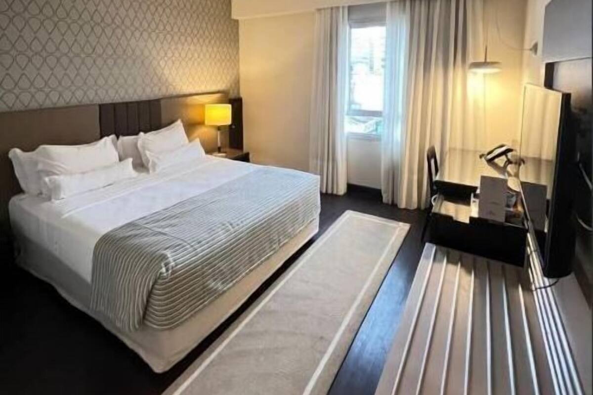 Habitación de hotel con una cama grande, un escritorio con silla, televisión y ventana con cortinas.