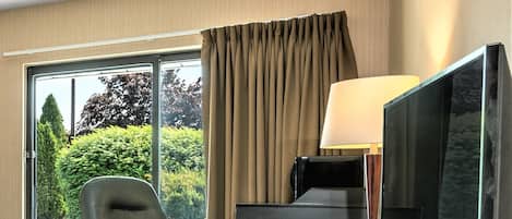 A hotel room with a desk, chair, TV, and large windows with curtains.