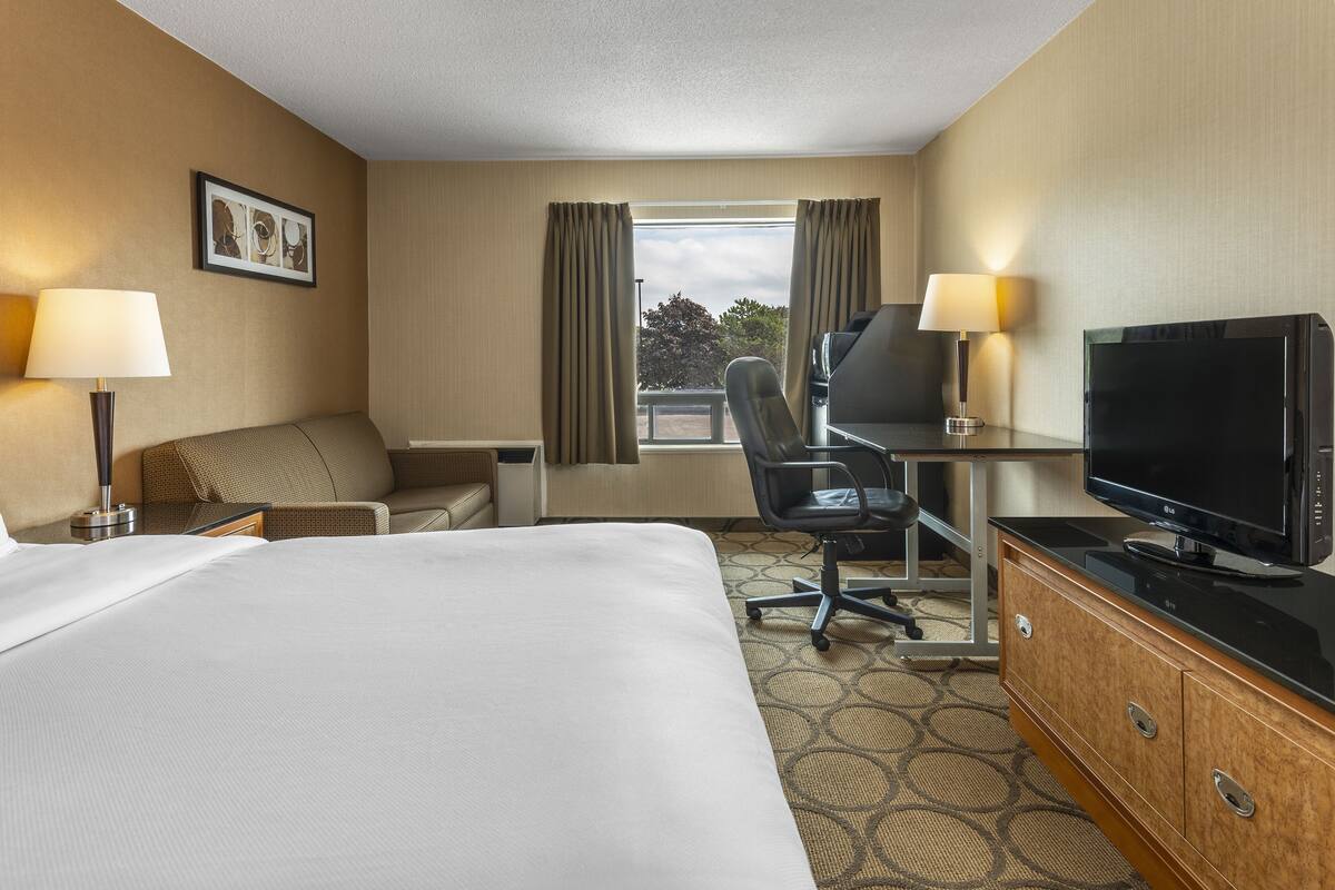 A hotel room with a large bed, a desk with a television, a chair, a sofa, and two lamps.